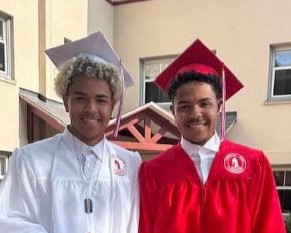 Alex and Anderson Dacruz, twin brothers with brown skin and short curly hair. Anderson has blonde hair and is wearing a white cap and gown. Alix has dark brown hair and is wearing a red cap and gown.