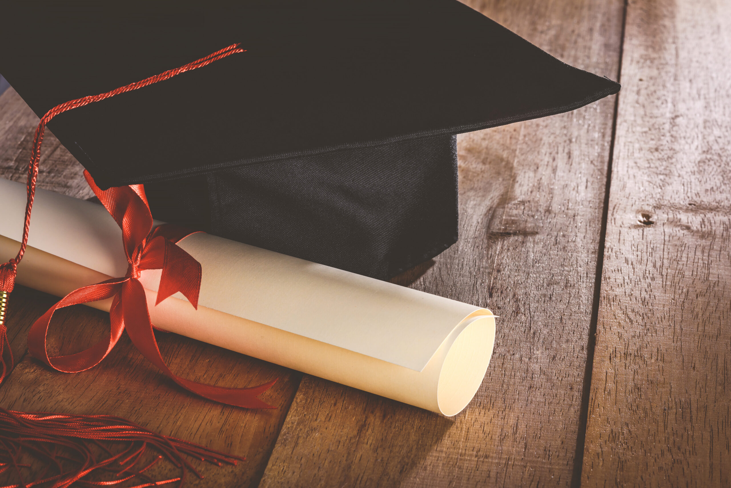 martarboard or graduation cap with red tassel and diploma tied with red ribbon on wooden table, vintage tone