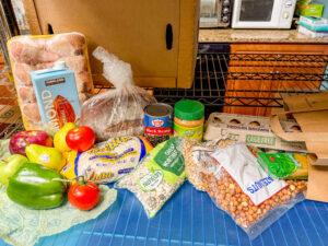 Example of an unpacked EPH food pantry grocery bag with a pack of chicken drumsticks, a loaf of bread, almond milk, a green pepper, tomatoes, apples, pears, rice, beans, nuts, peanut butter, and eggs.