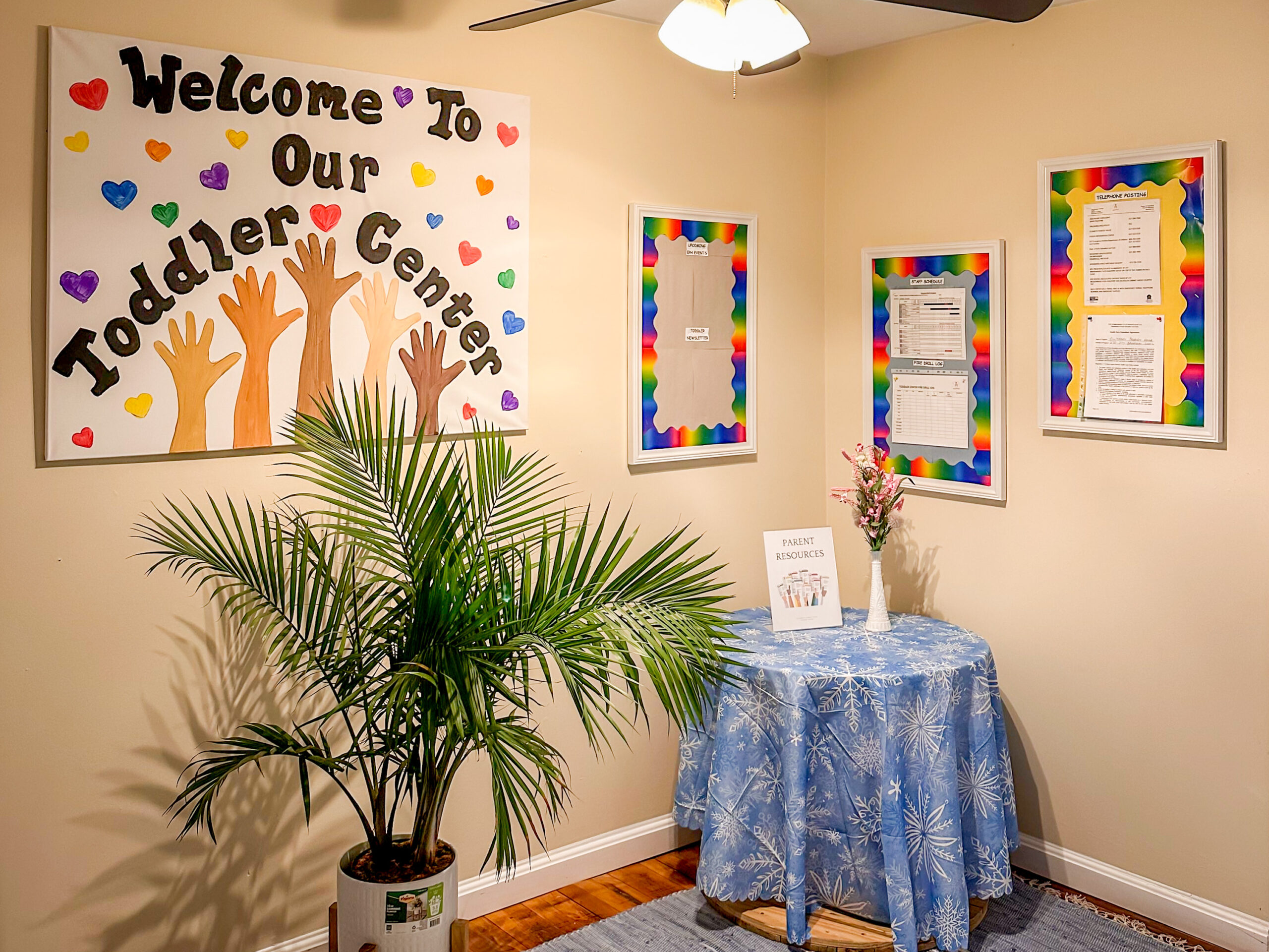 A white sign that reads "Welcome to Our Toddler Center" with multicolor hearts & hands hangs on a warm wall above a potted palm frond and a small round table covered by a blue cloth.