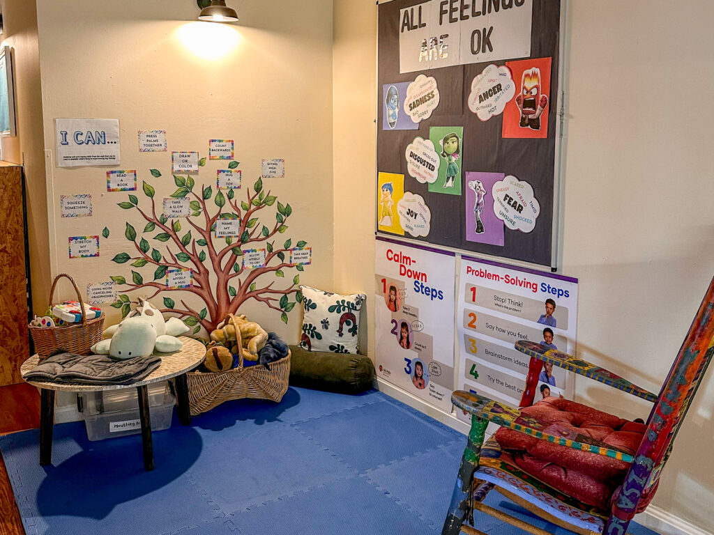 A room with a mural of a tree on one wall and a bulletin board that reads "all feelings are okay" on the other. The floor is covered in blue foam tiles. The room includes a rocking chair, a short round table with books and a basket on it, a larger basket sits nearby with pillows and plush toys.