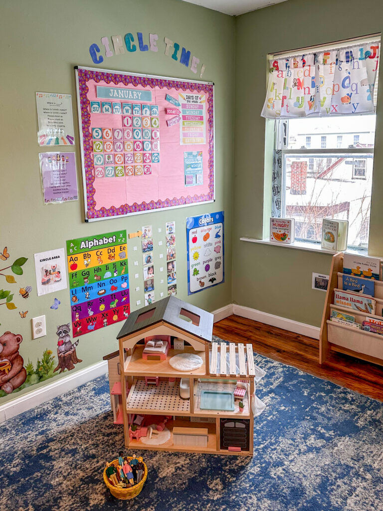 A room with green walls and hardwood floors covered by a blue and white rug. The room features a large window, a large bulletin board titled "circle time" and other posters. There is a wooden dollhouse on the rug and a low bookshelf against the wall with the window. 